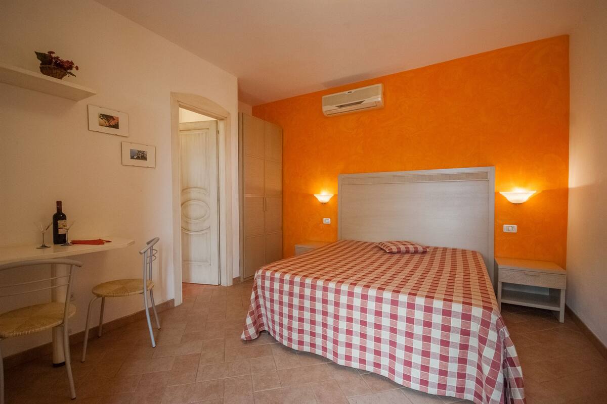 Une chambre avec une couvre-lit à carreaux, un bureau avec une chaise, une bouteille de vin et deux lampes fixées au mur.