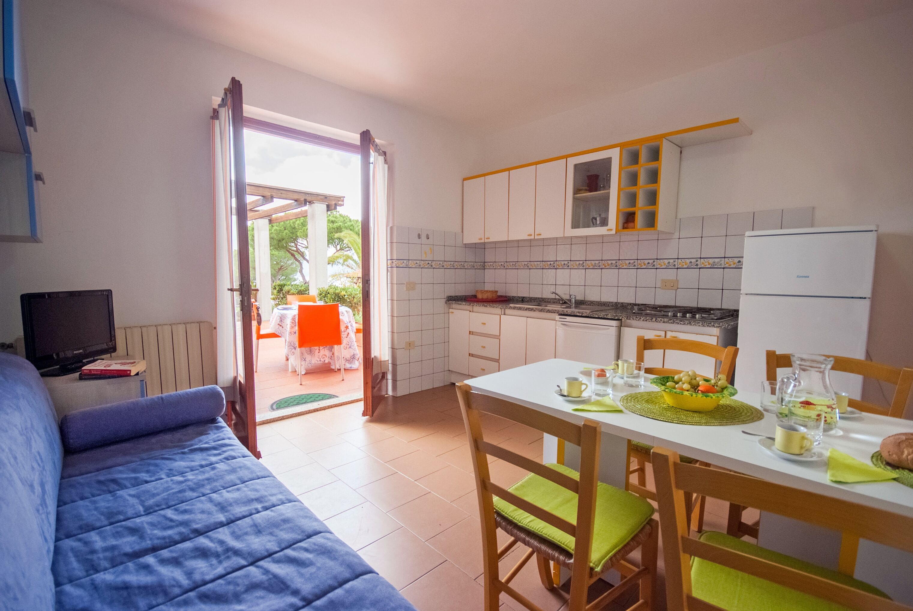 Cocina compacta con gabinetes blancos, una mesa de comedor preparada para cuatro personas y un televisor sobre un mueble.