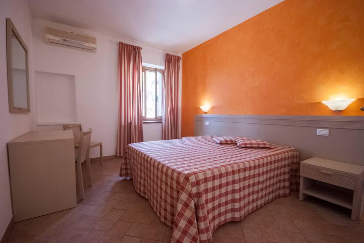 Une chambre à coucher avec une couvre-lit à carreaux, un bureau, une chaise, une table de chevet et une lampe fixée au mur.