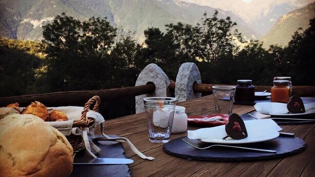 Una mesa de madera dispuesta con una cesta de pan, un vaso de agua, un frasco de mermelada y un plato con un postre de chocolate, con vistas a un paisaje montañoso.