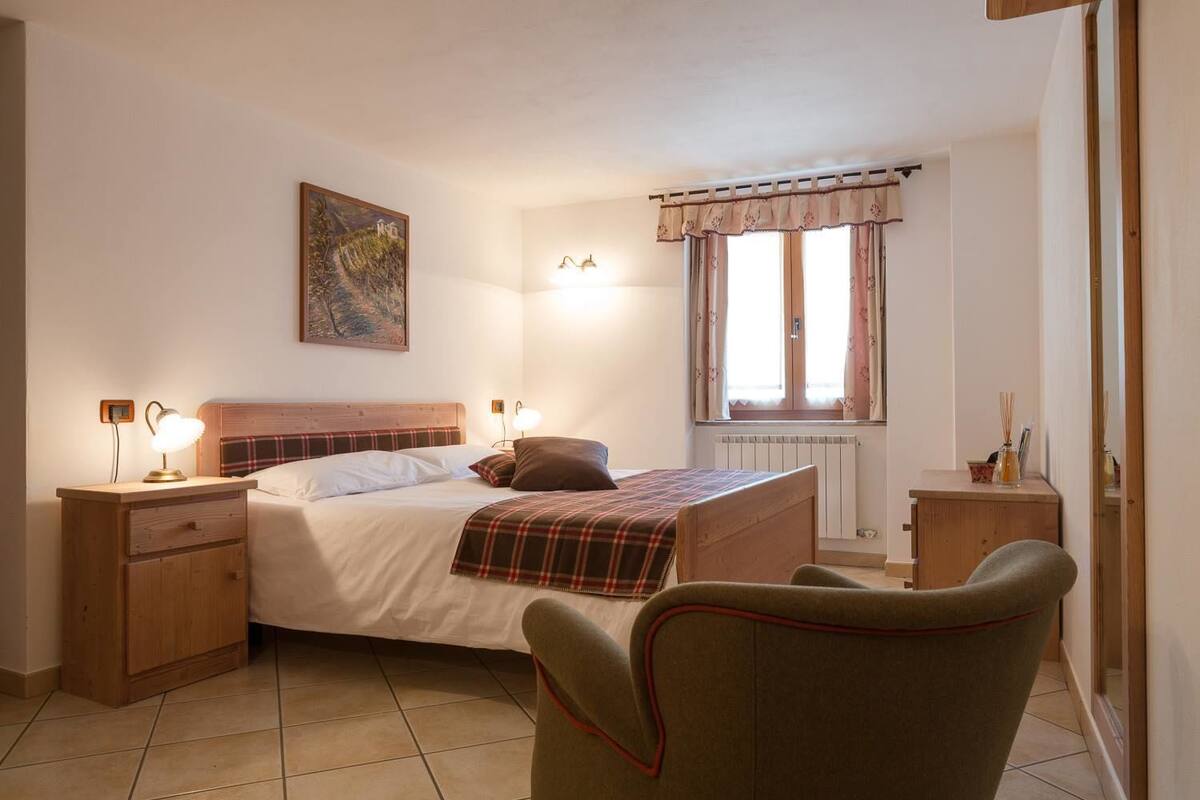 Un dormitorio con cama, mesitas de noche, una silla, una ventana con cortinas, un radiador y un cuadro en la pared.