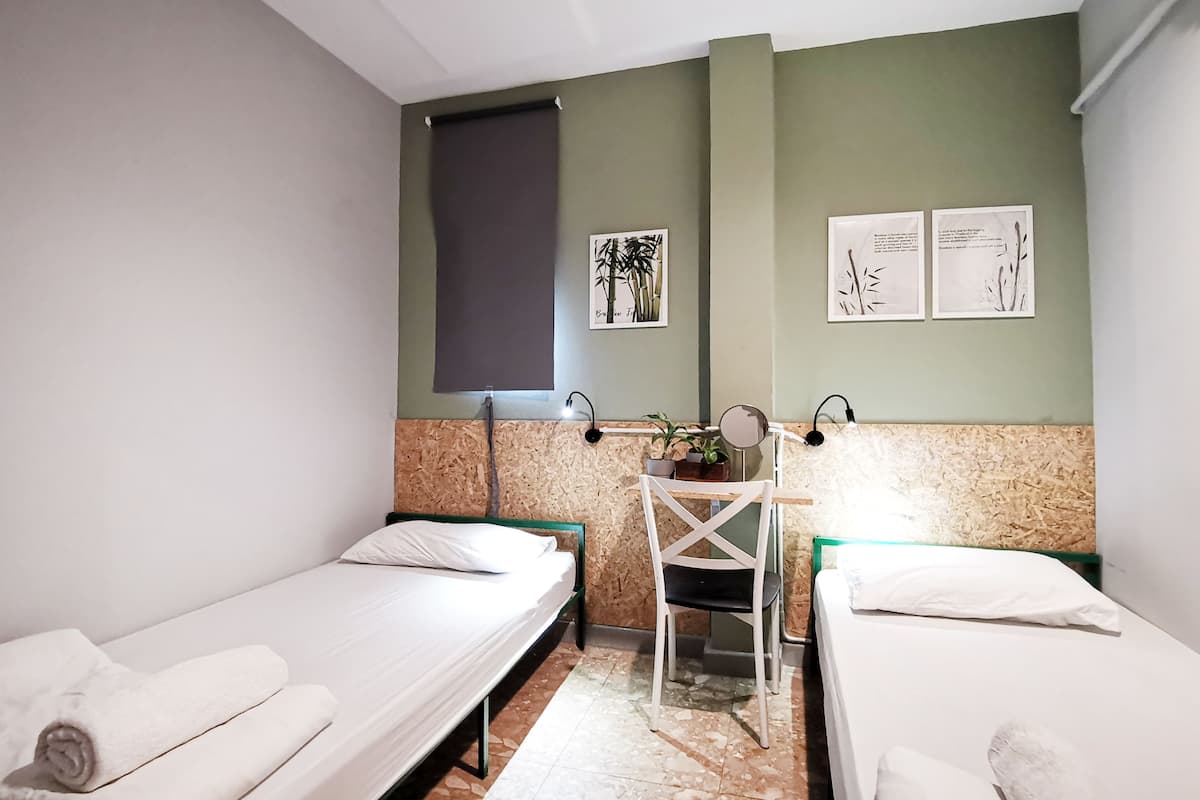 Ein kleines, sauberes Zimmer mit zwei Einzelbetten, einem Schreibtisch mit Stuhl und einem Fenster mit Jalousie.
