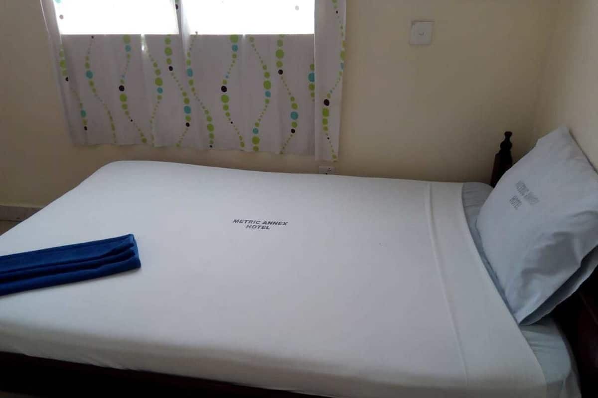 A single bed with a white mattress and a blue towel, a white pillow, and a wall-mounted light switch.