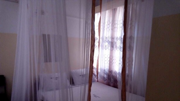 A room with a bed, a bedside table, and a window with sheer curtains. - Metric Annex Hotel (Mombasa)
