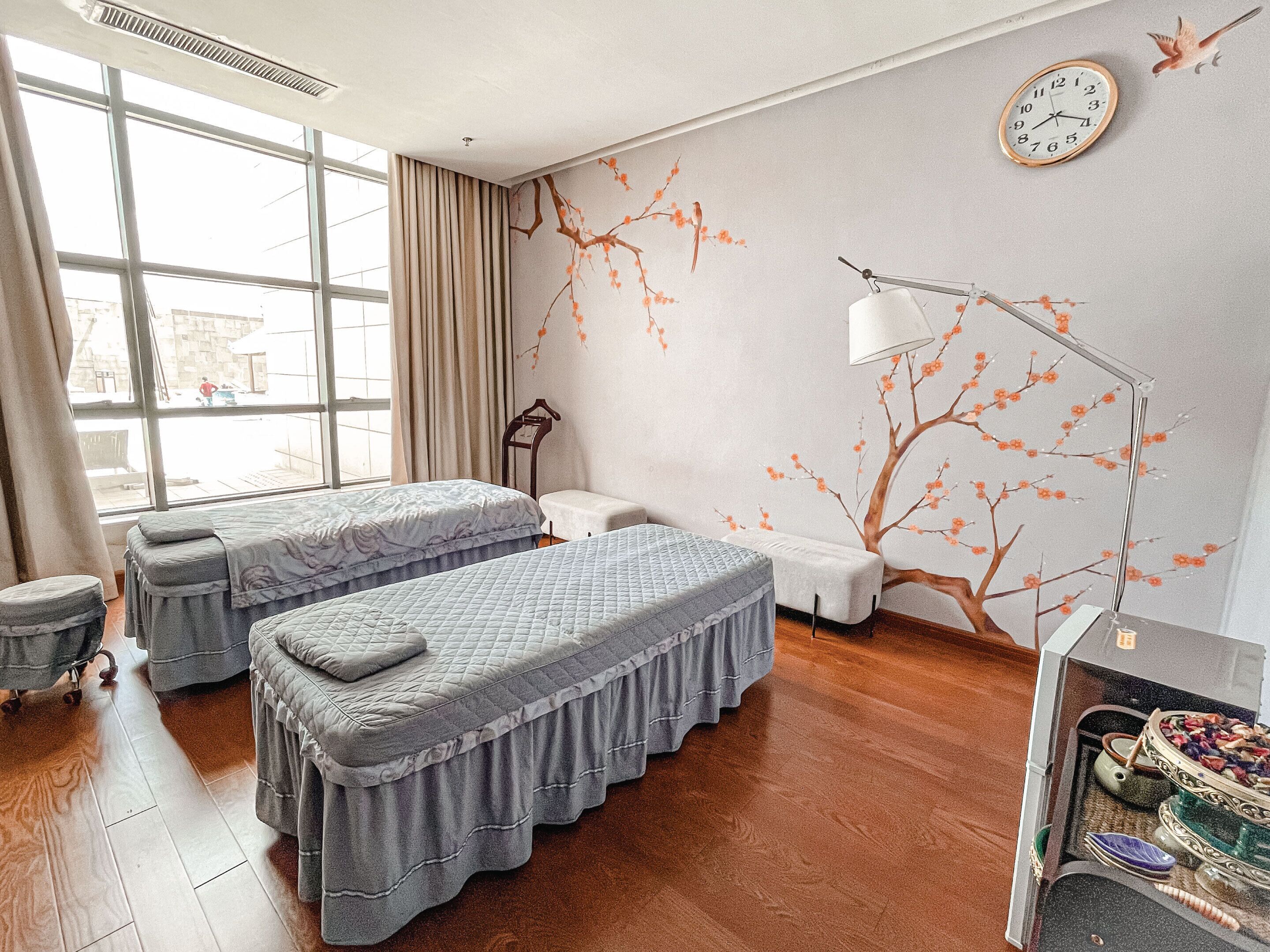 A hotel room with two beds, a large window, a wall clock, and a decorative tree mural.