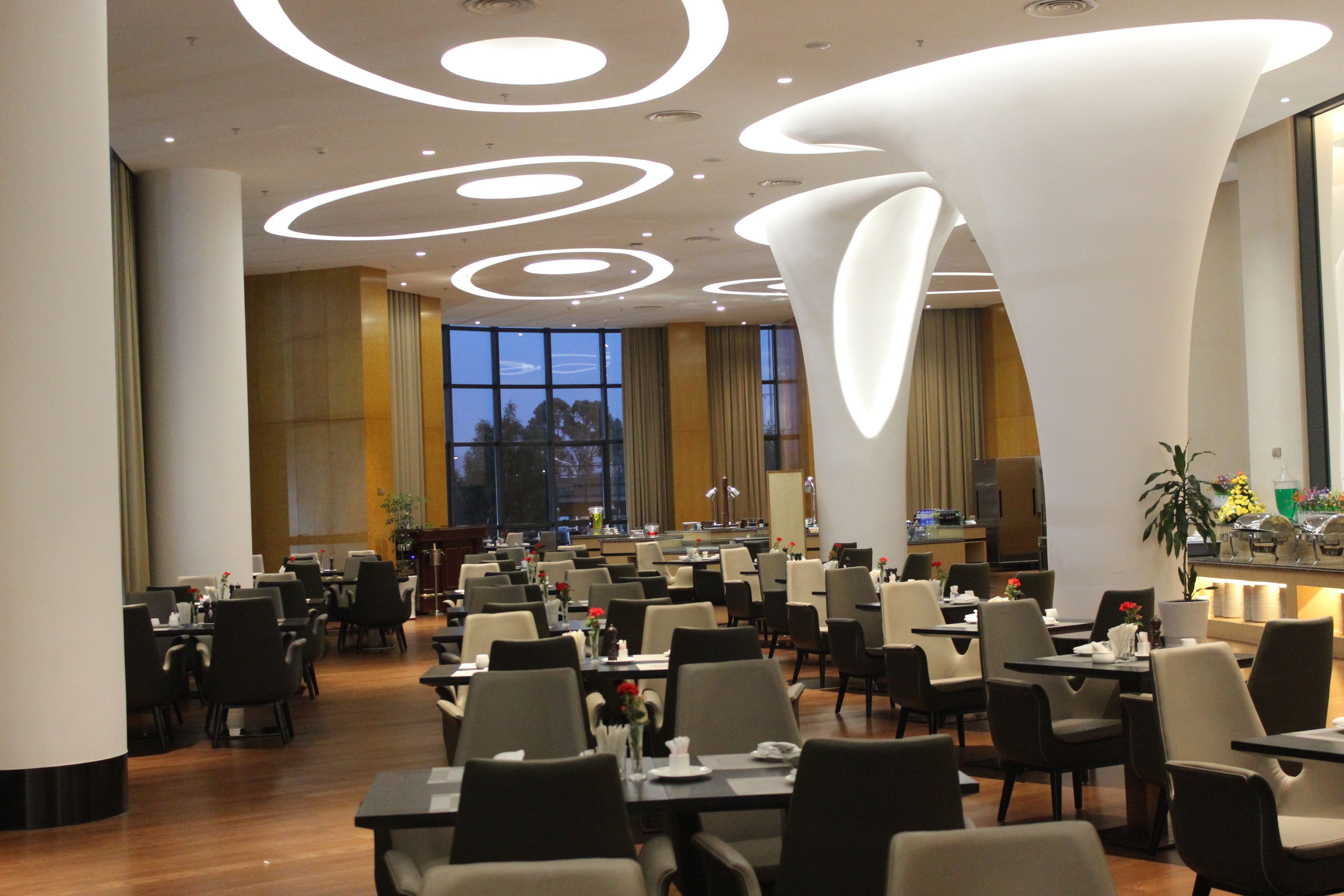 A spacious dining area with multiple tables and chairs, modern ceiling lighting, and large windows providing natural light.