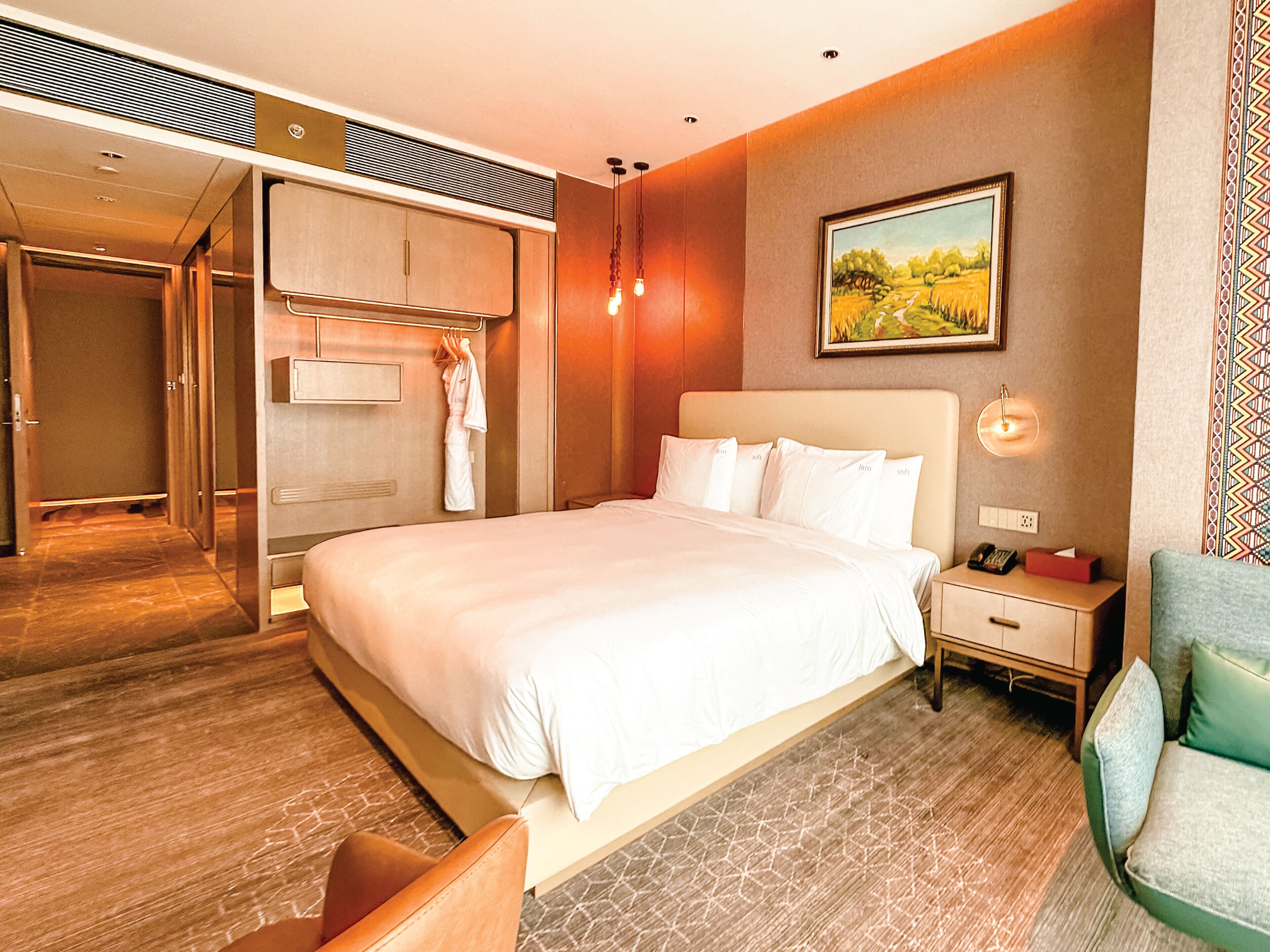 A hotel room with a large bed, a bedside table with a lamp, a painting on the wall, and a wardrobe.