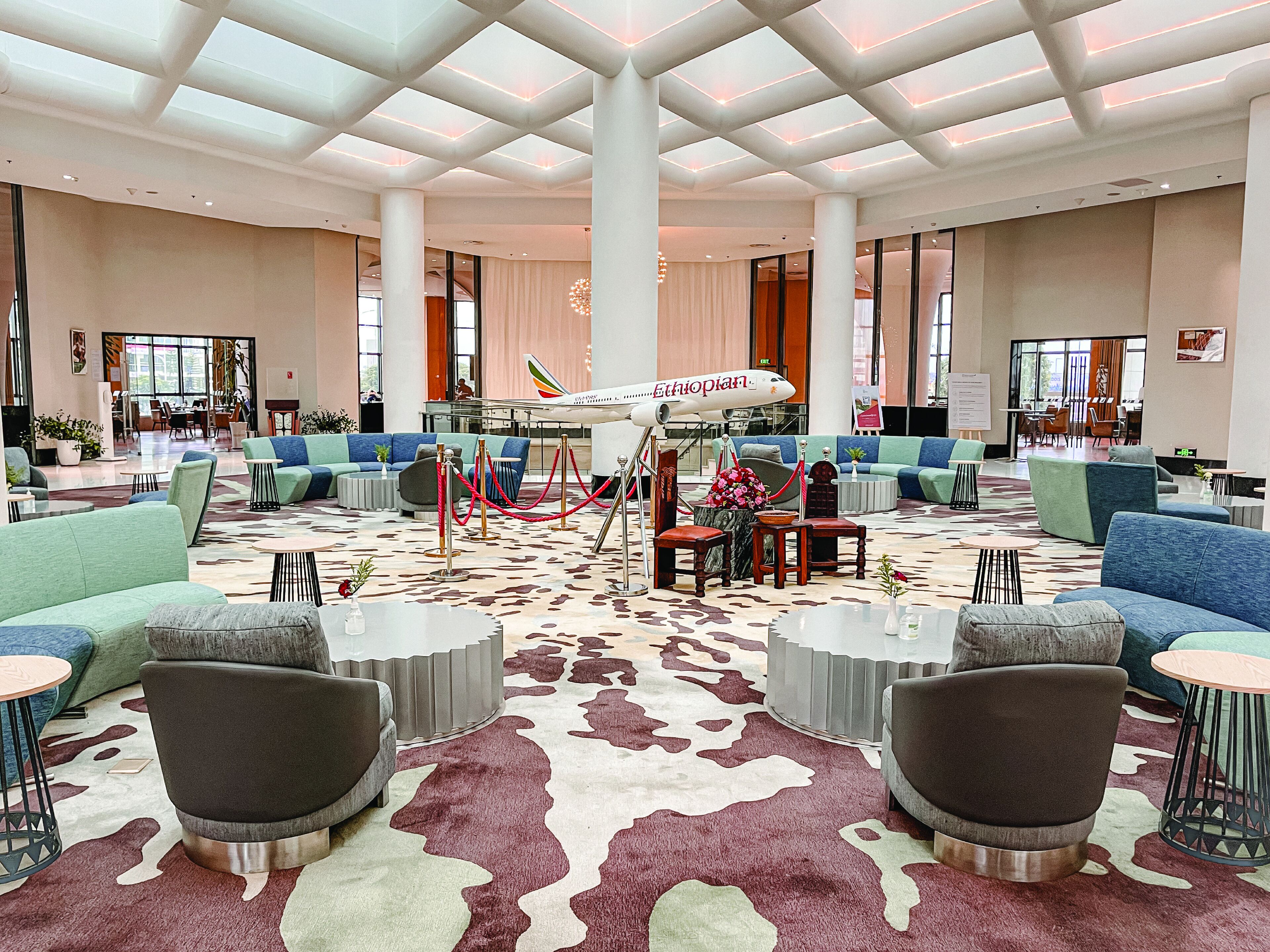 A spacious lobby with a modern design, featuring a central seating area with sofas and armchairs, a patterned carpet, and a large Ethiopian Airways airplane model as a centerpiece.