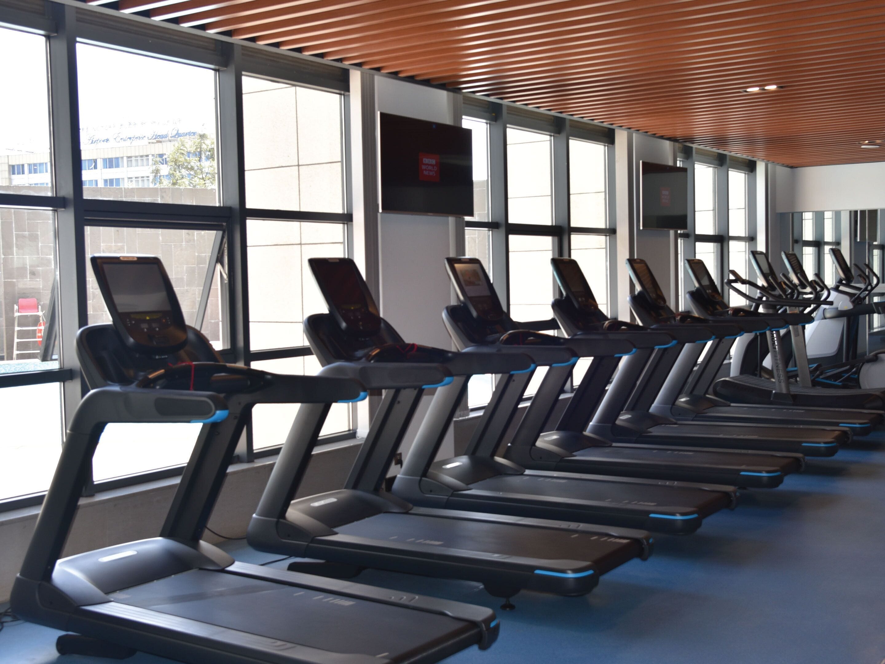 A row of treadmills in a gym with large windows and a wooden ceiling.