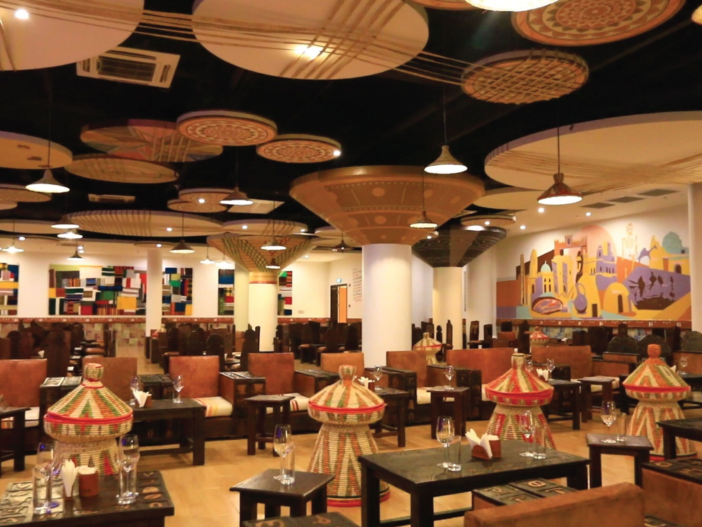 Een restaurant met tafels, stoelen, hanglampen en een muurschildering.