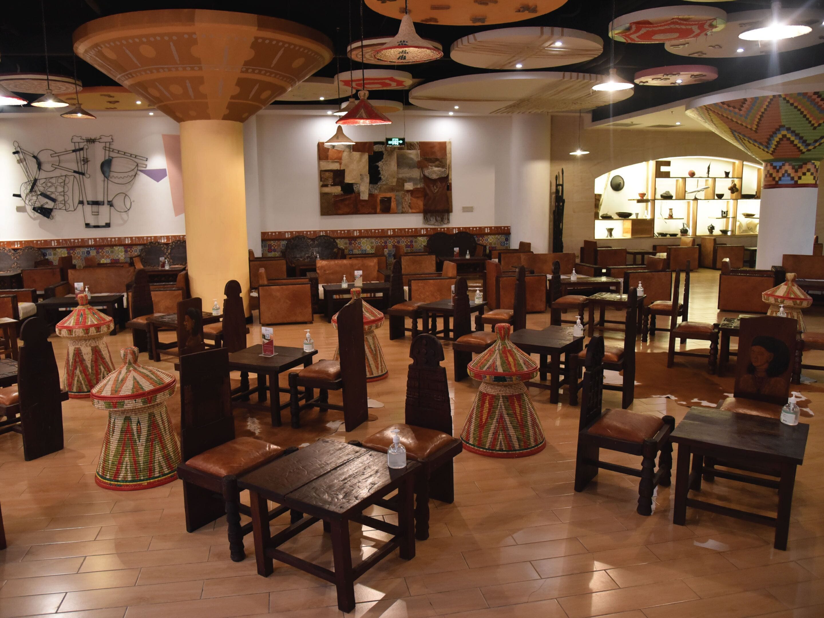 A restaurant with wooden tables and chairs, decorative hanging lamps, and a tiled wall with artwork.
