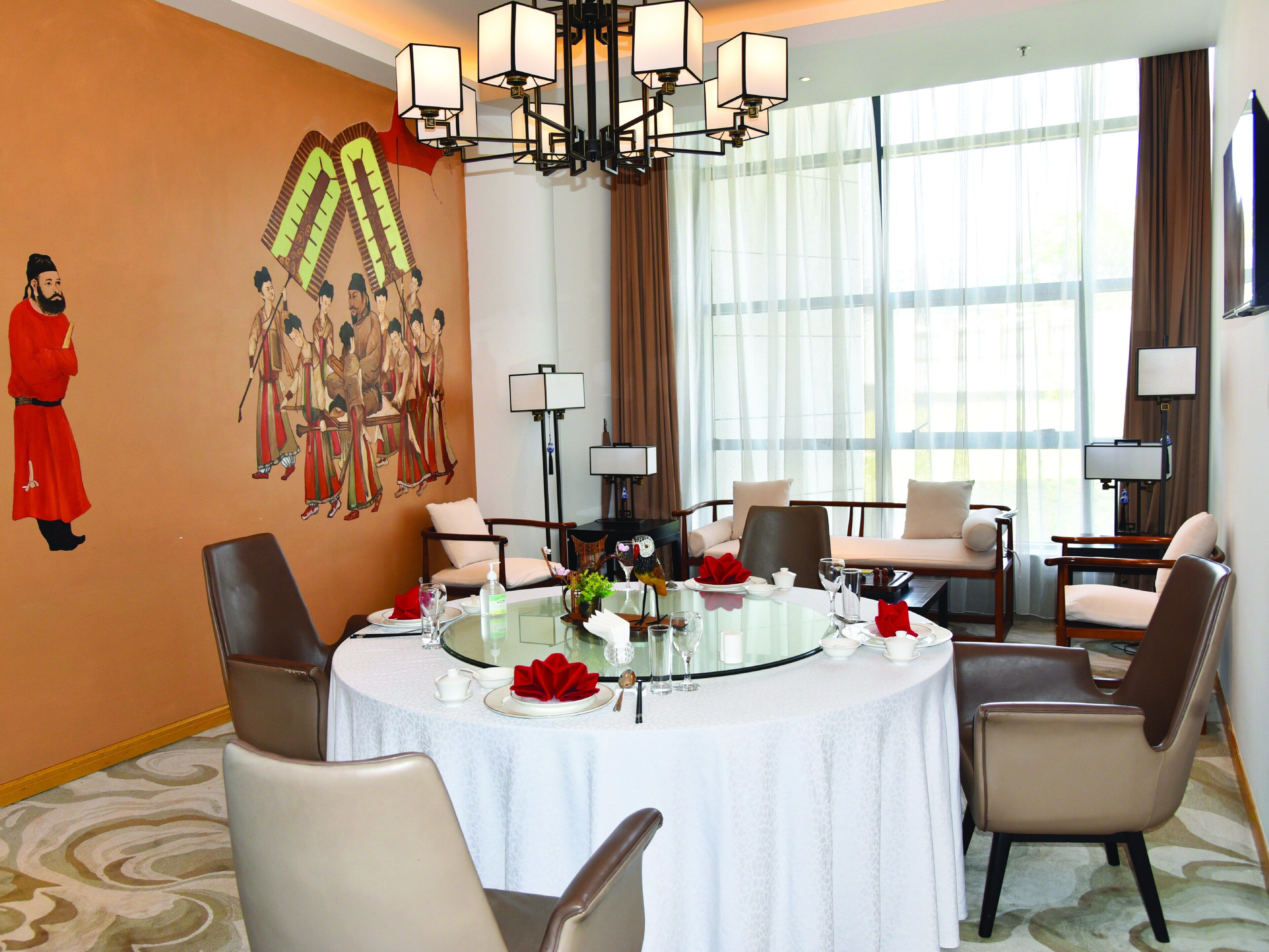 A hotel dining area with a round table set for four, surrounded by chairs, a chandelier, and a mural on the wall.