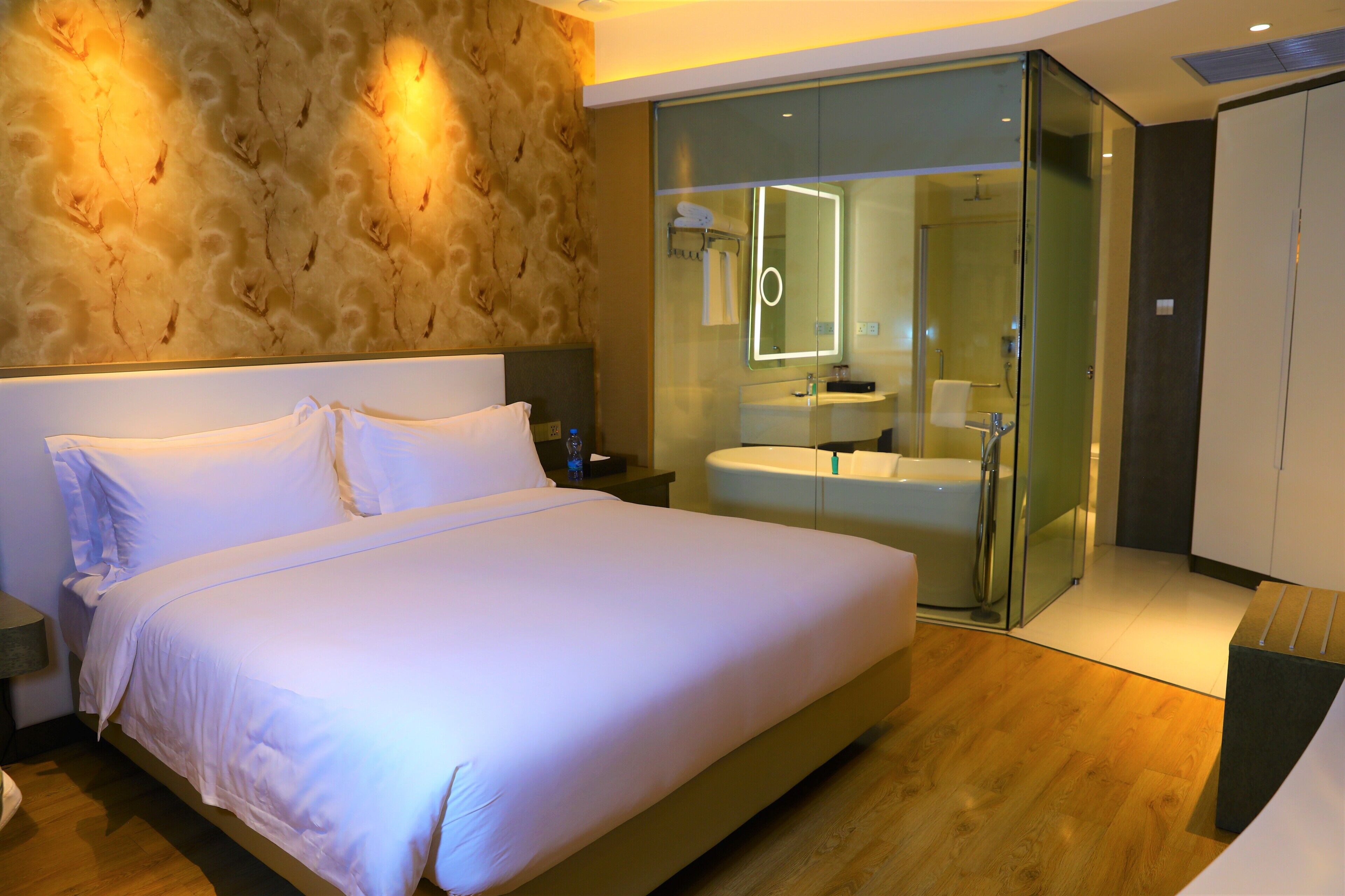 A modern hotel room with a large bed, a glass-enclosed bathroom, and a wooden floor.