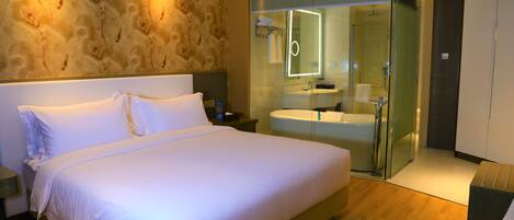 Quarto de hotel moderno com uma cama grande, banheiro envidraçado e piso de madeira.