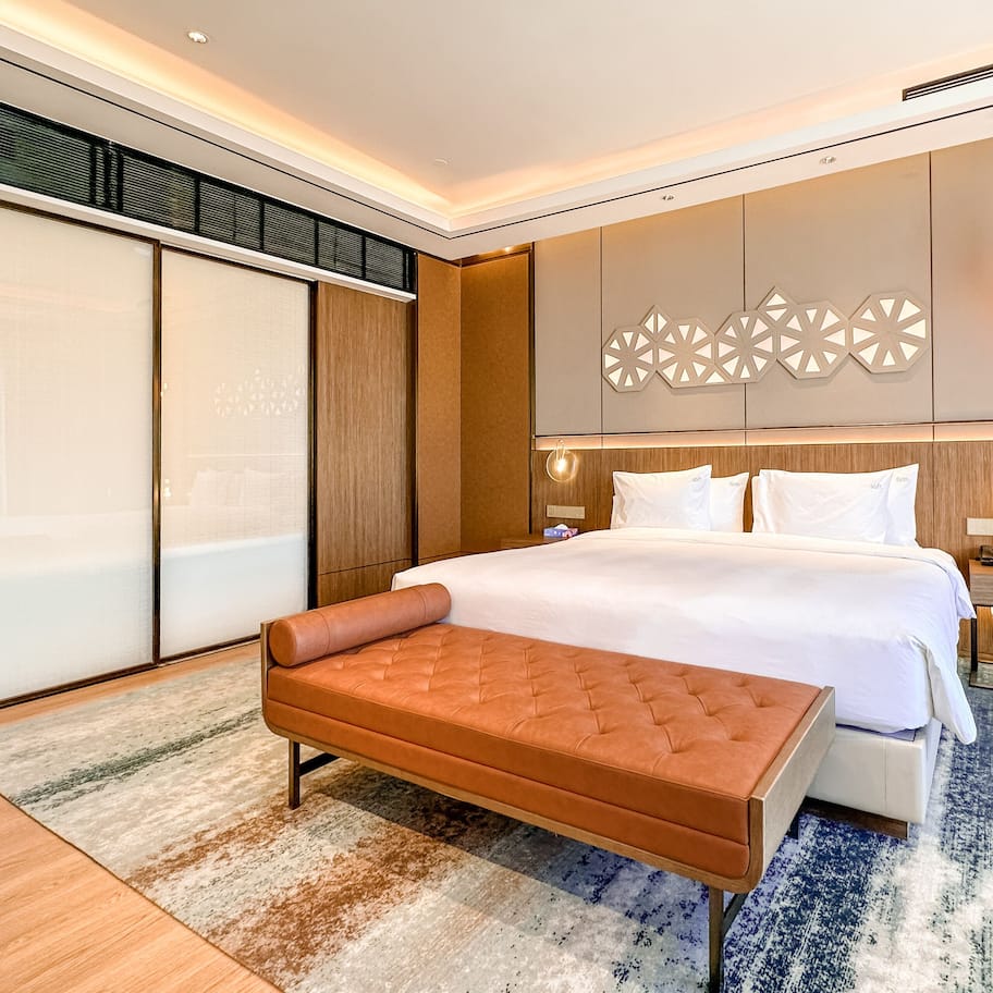 A modern hotel room with a large bed, a bench, a bedside table, and a wall-mounted light fixture.