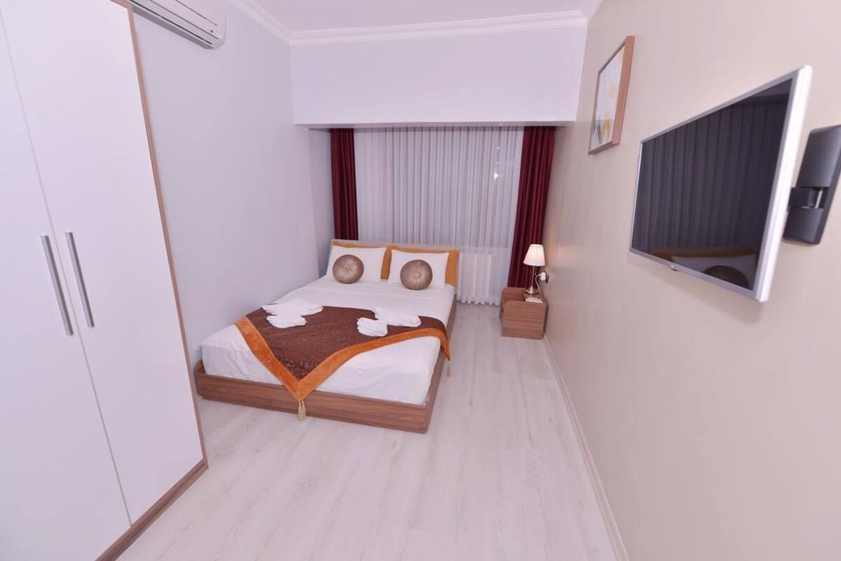 Une chambre moderne avec un lit en bois, des murs blancs, une grande fenêtre avec des rideaux, une table de chevet avec une lampe et un téléviseur fixé au mur.