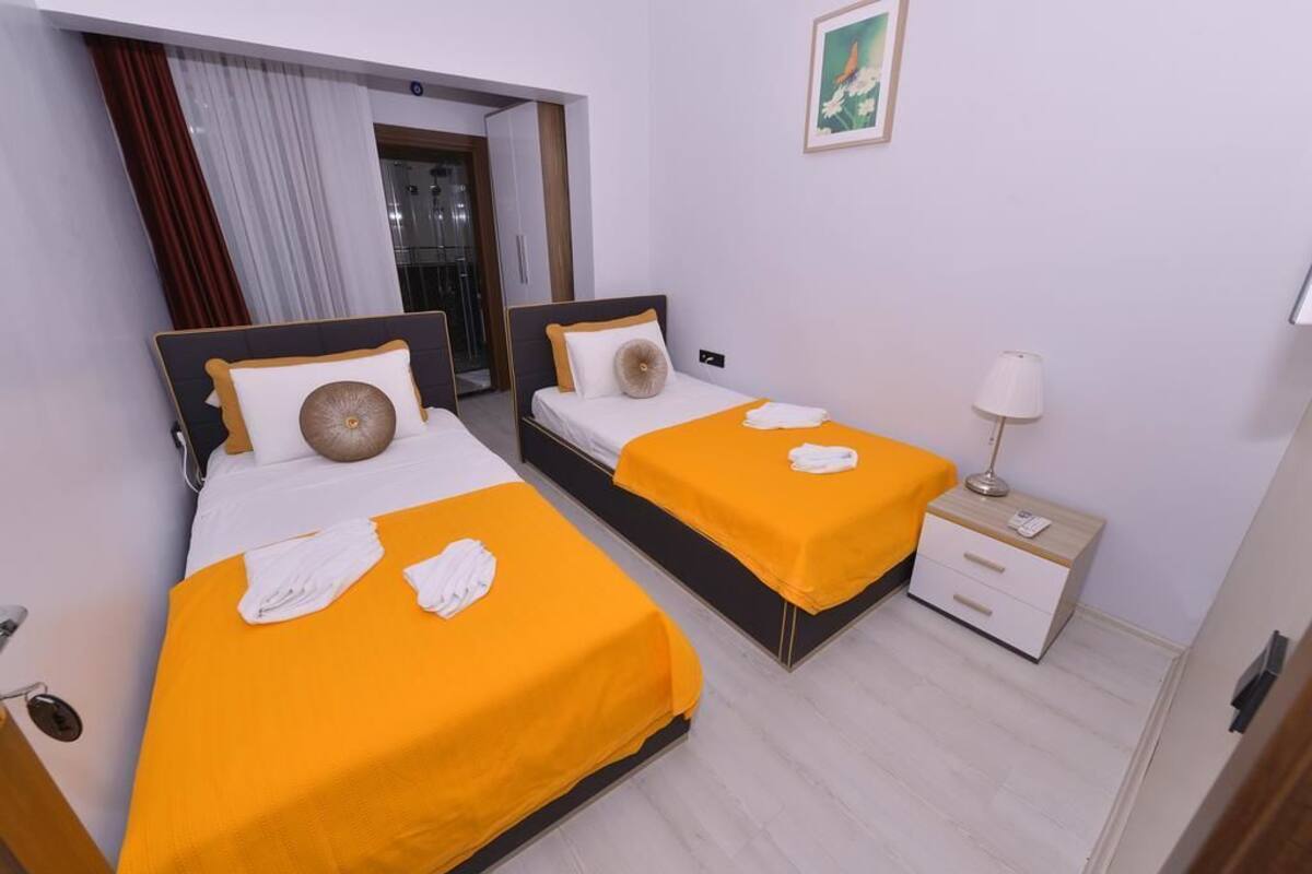 Une chambre d’hôtel avec deux lits, chacun recouvert d’une couverture orange et agrémenté d’oreillers blancs. Il y a une petite table de chevet avec une lampe et un tiroir. Les murs de la chambre sont blancs et il y a une fenêtre avec des rideaux.
