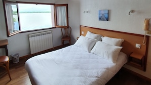 Una cama bien tendida con sábanas blancas, un cabecero de madera, una mesita de noche con una lámpara, una silla y una ventana con vista a un cuerpo de agua.