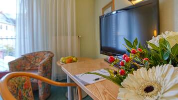 Habitación de hotel con un escritorio de madera, televisor, una butaca floral y un arreglo de flores.