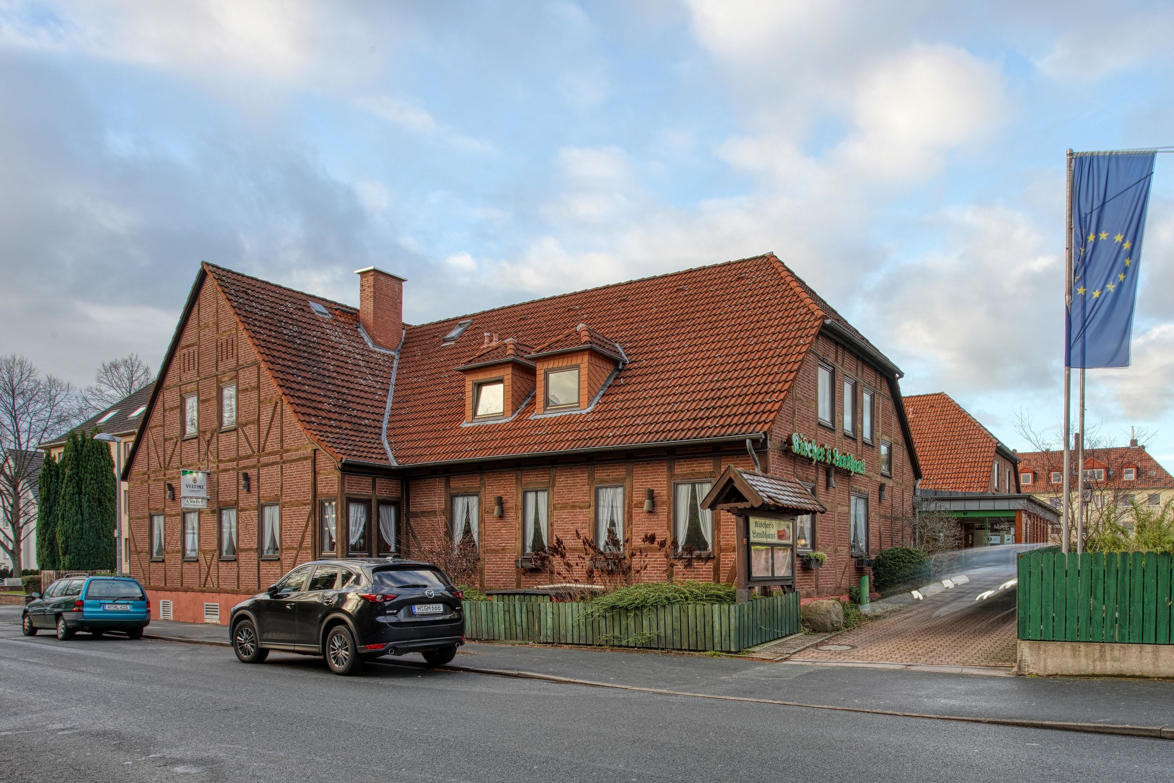 Un bâtiment traditionnel en briques, doté d’un toit en pente, d’une entrée bien visible et d’un drapeau qui laisse supposer qu’il pourrait s’agir d’un hôtel.