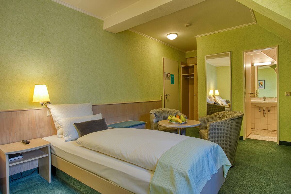 A hotel room with a bed, bedside table, lamp, chair, and a small table with fruits.