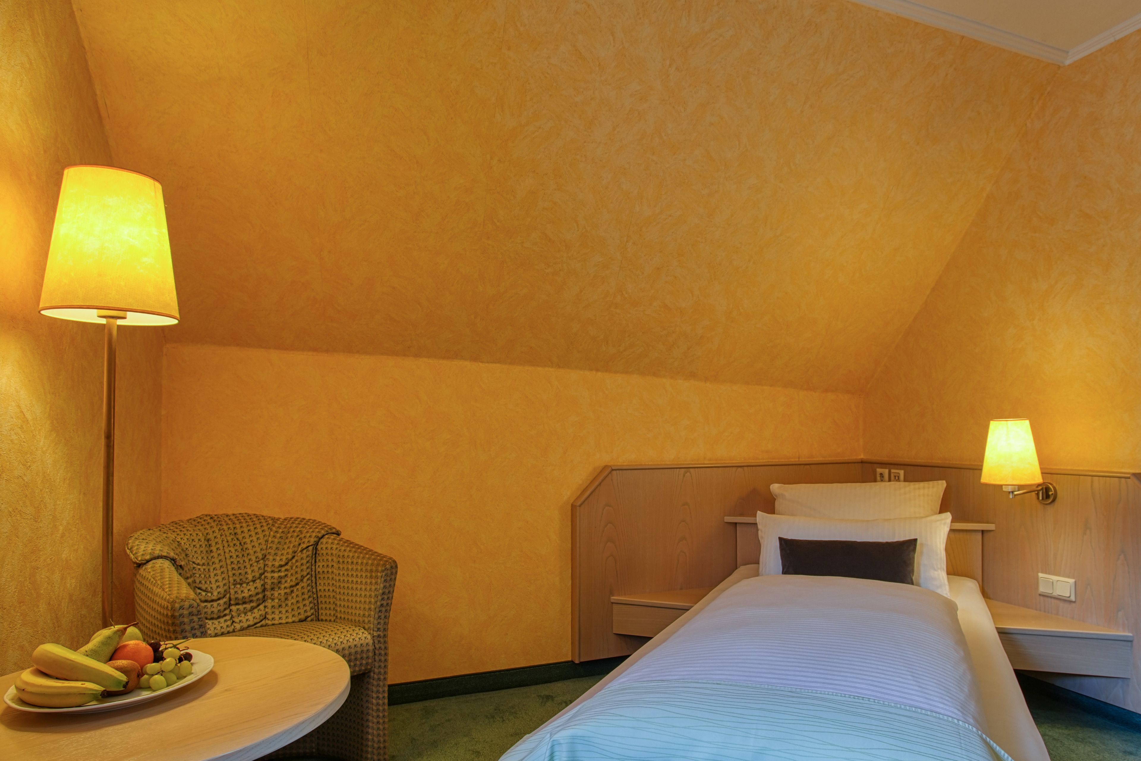 A hotel room with a bed, a small round table with fruit, a chair, and two wall-mounted lamps.