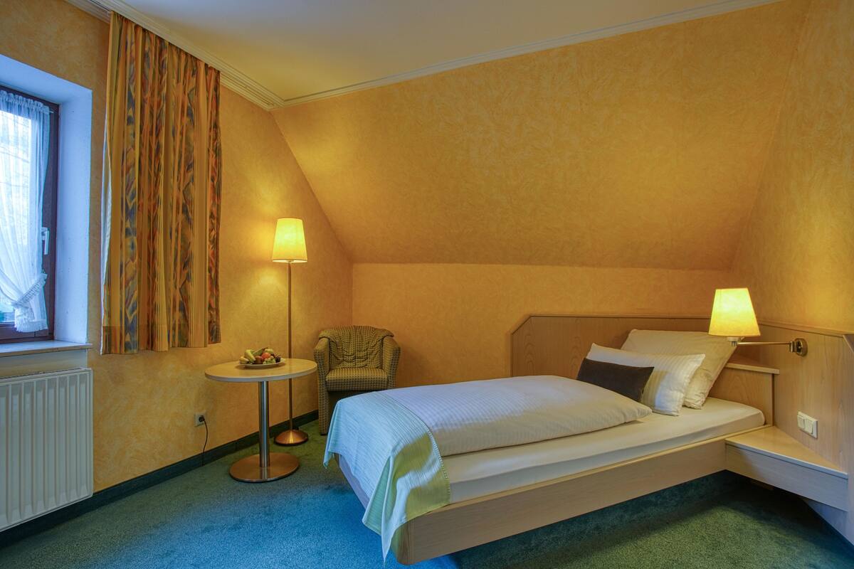 A hotel room with a single bed, bedside lamps, a chair, a small round table, and a window with curtains.