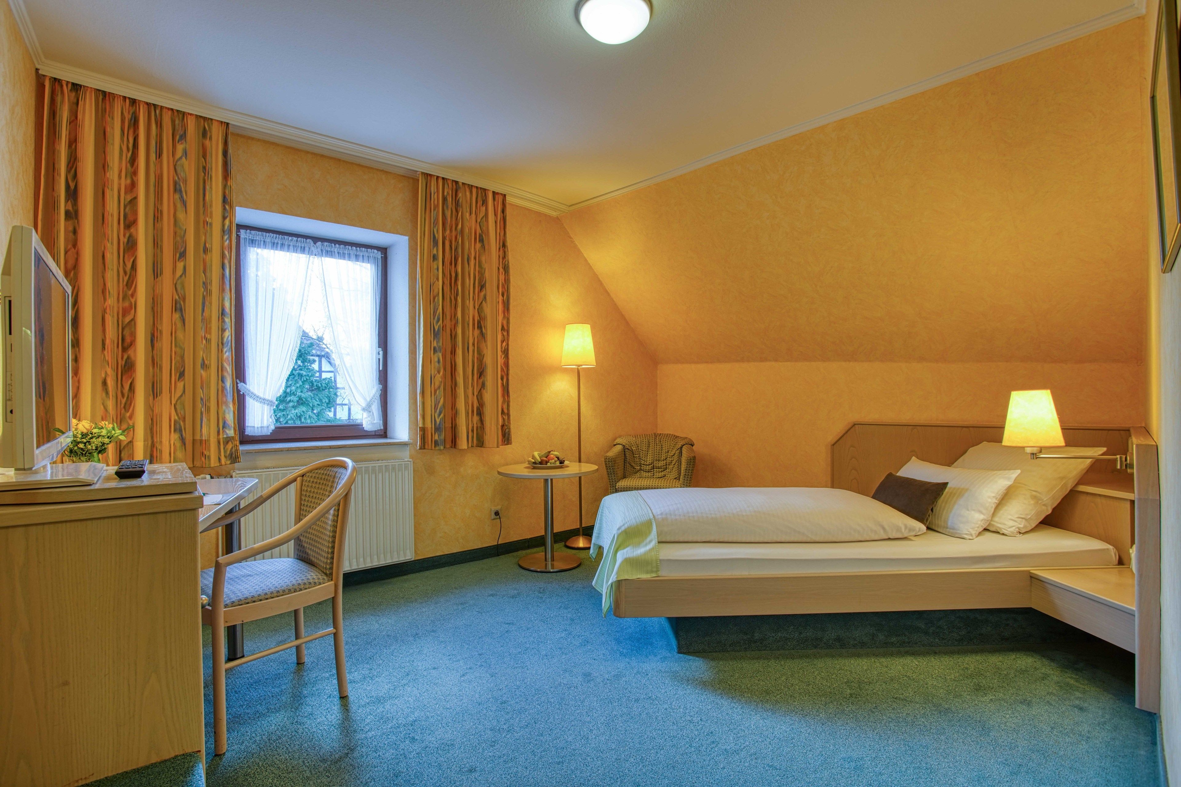 Een hotelkamer met een bed, een bureau, een stoel, een lamp en een raam met gordijnen.