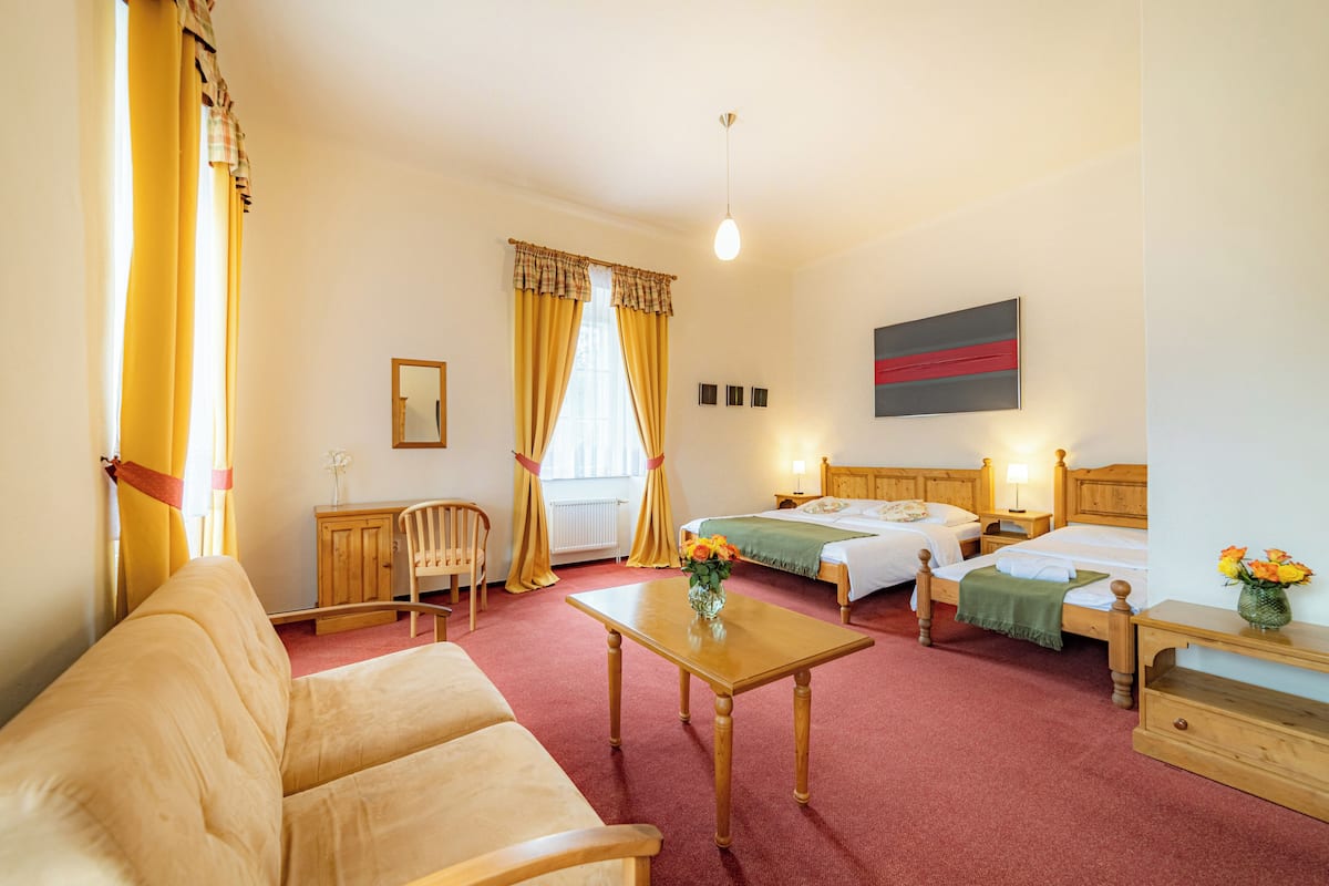 Quarto de hotel com cama e escrivaninha de madeira, uma cadeira, um sofá e uma janela com cortinas.