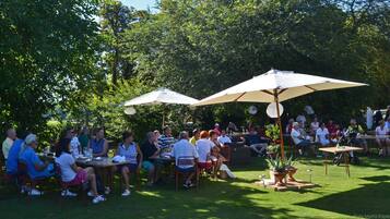An outdoor gathering with people seated at tables under umbrellas, surrounded by greenery.