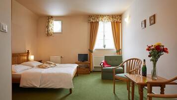 A hotel room with a bed, a TV, a chair, a table with a bottle and flowers, and a window with curtains.
