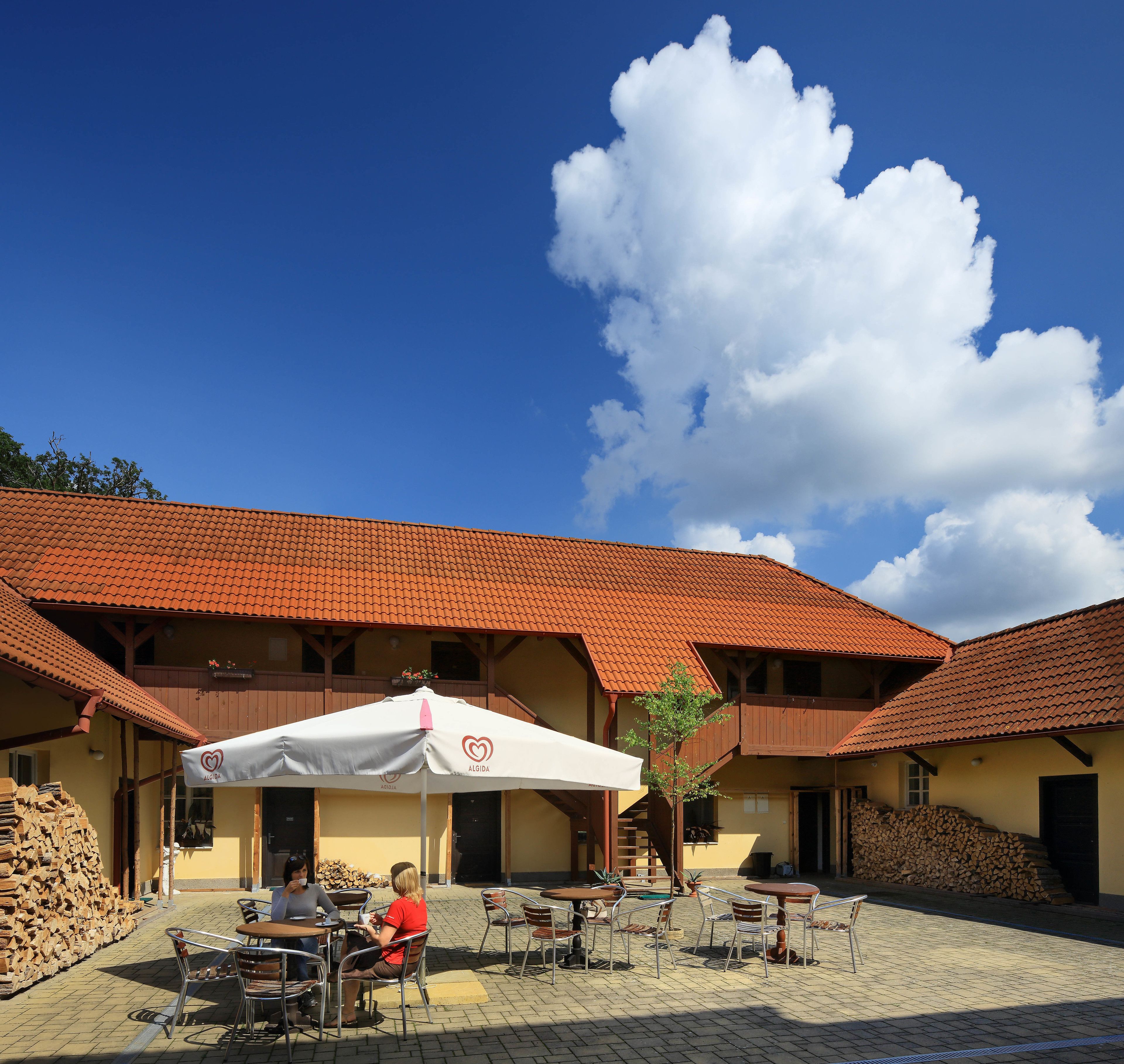 Ein Außenbereich mit Tischen und Stühlen, ein Gebäude mit einem Ziegeldach und ein klarer, blauer Himmel mit Wolken.
