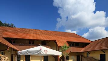 Ein Außenbereich mit Tischen und Stühlen, ein Gebäude mit einem Ziegeldach und ein klarer, blauer Himmel mit Wolken.