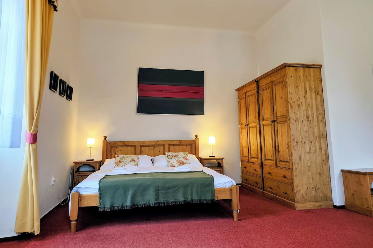 Quarto com cama, abajures, guarda-roupa de madeira, uma obra de arte na parede e janela com cortinas.