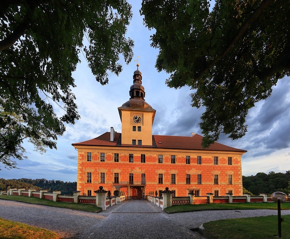A large, historic building with a prominent clock tower, a symmetrical facade, and a paved entrance area.