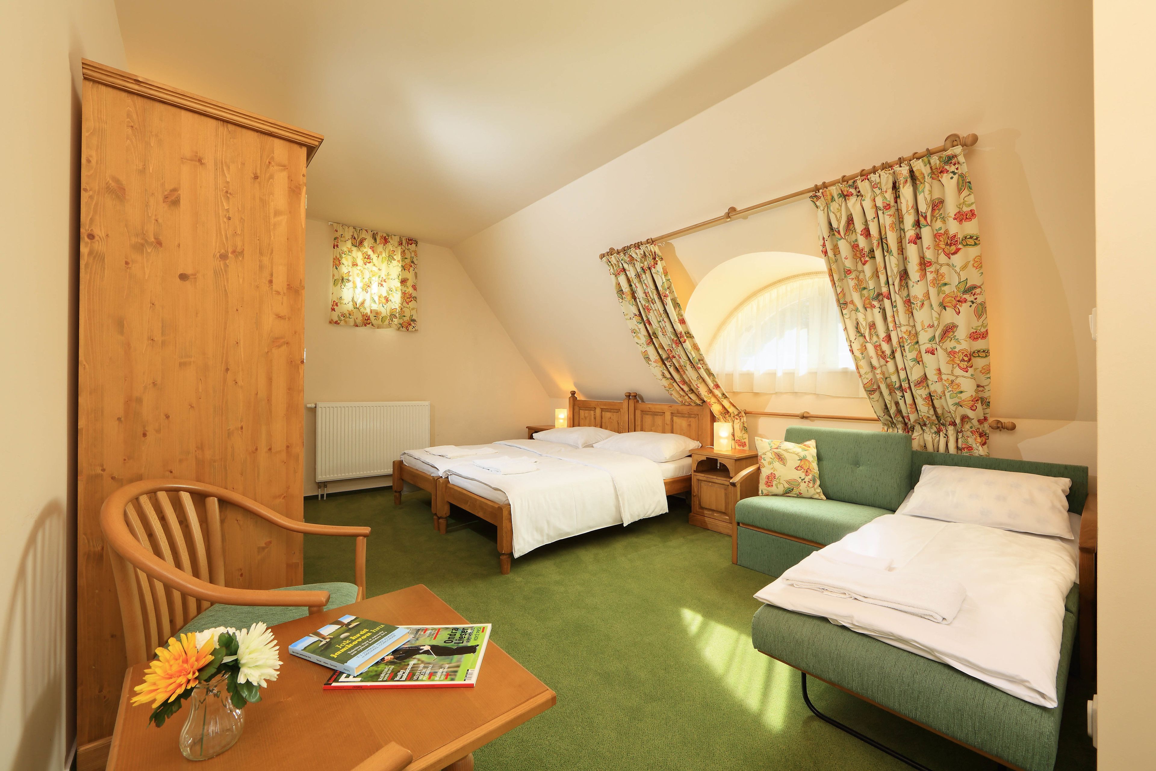 Un dormitorio con dos camas, un armario de madera, un sofá verde, una mesa con un libro y flores, y una ventana con cortinas florales.