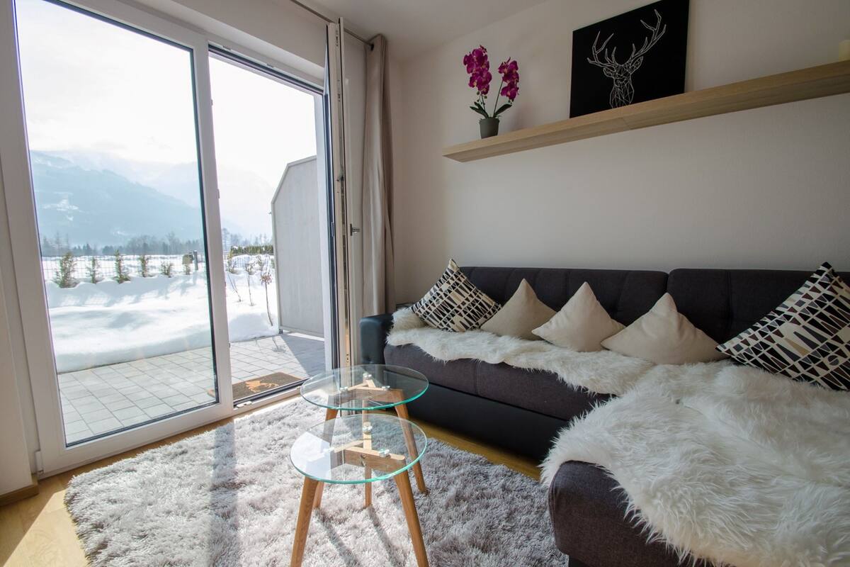 A modern living room with a sofa, glass coffee table, and a view of snow-covered mountains through a sliding glass door.
