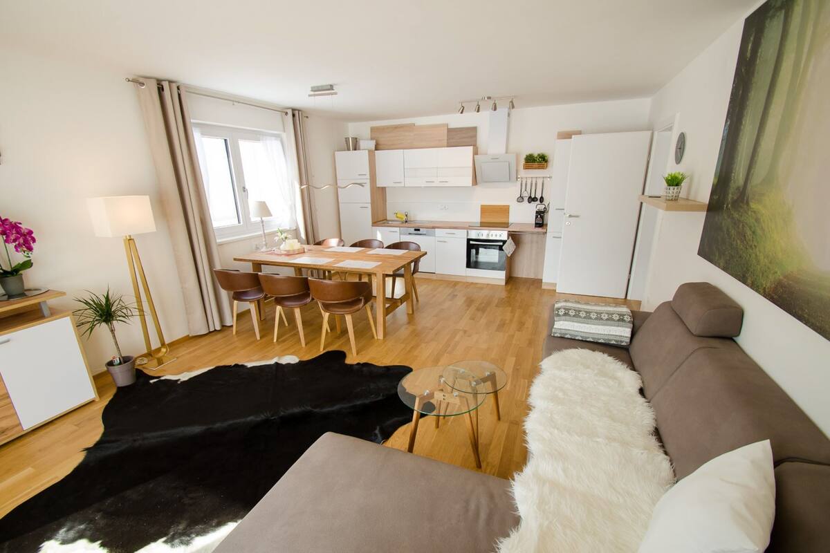 A modern living room with a sofa, a dining table with chairs, a kitchen area with cabinets and appliances, and a large window with curtains.