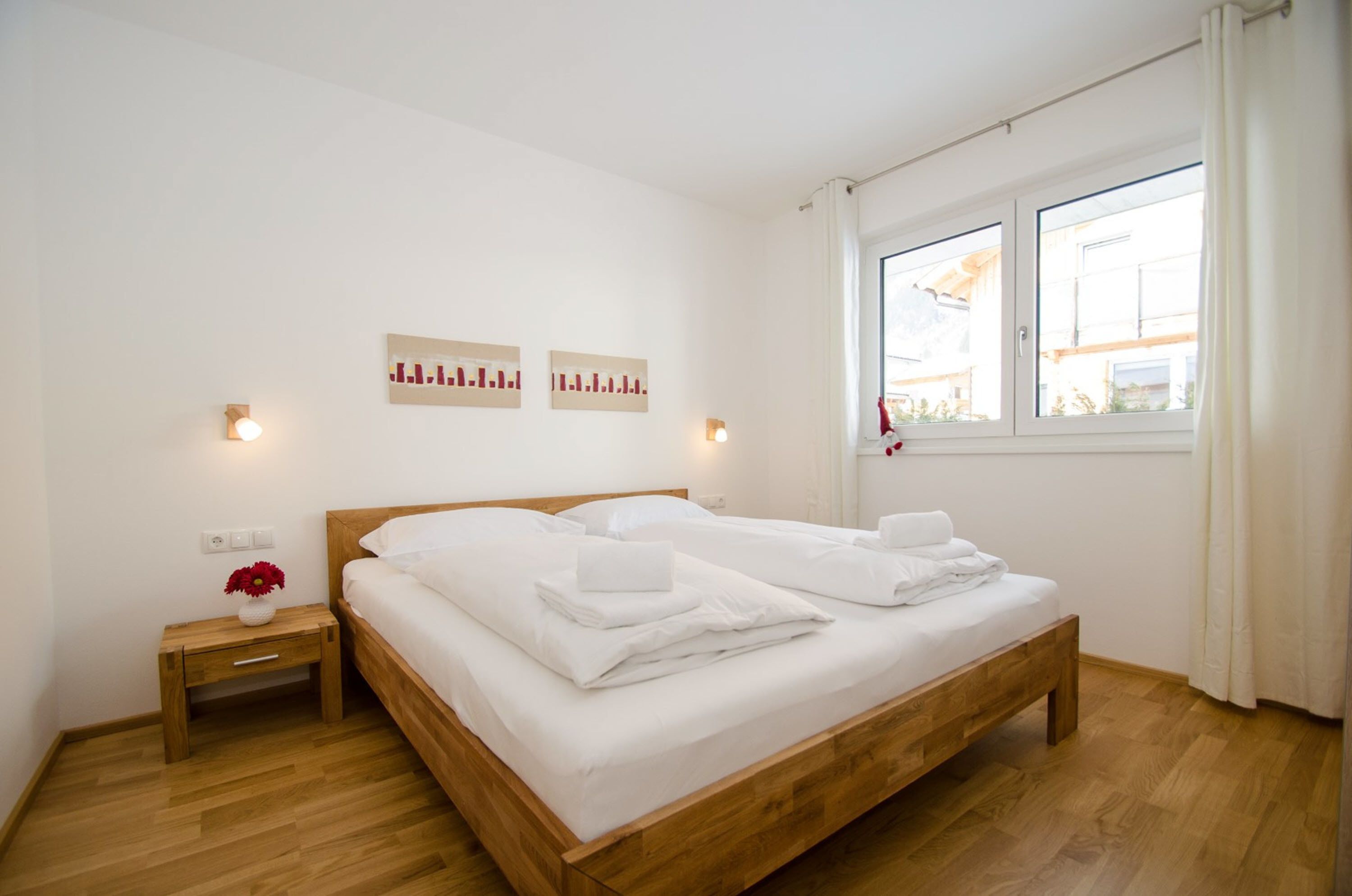 Ein ordentlich bezogenes Bett in einem Zimmer mit Holzboden, einem kleinen Nachttisch mit Blumenvase und zwei Wandleuchten.
