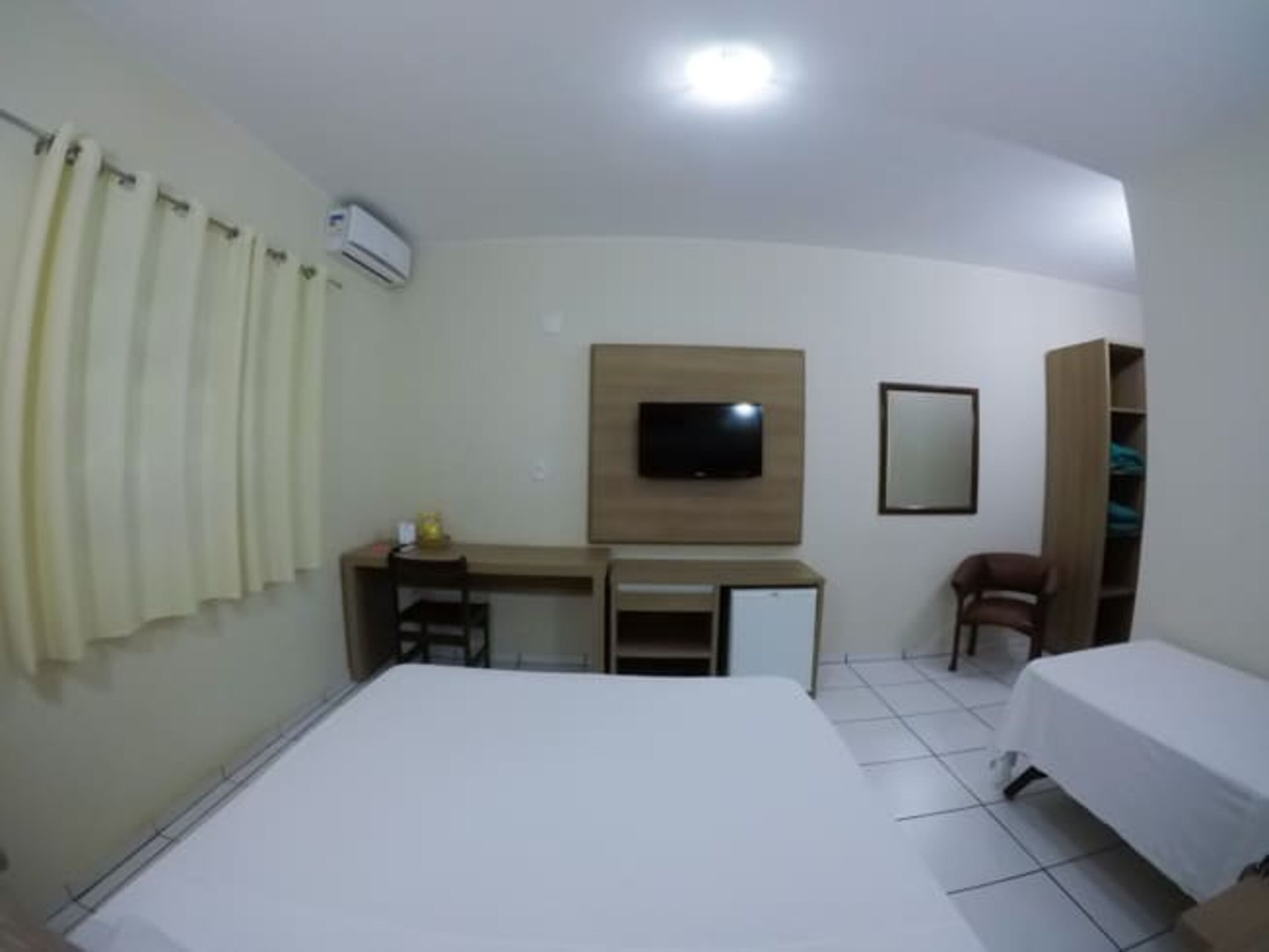 A hotel room with a bed, a desk with a chair, a television mounted on the wall, and a wardrobe.