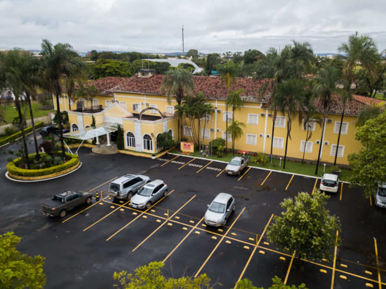 Un estacionamiento con varios coches, un edificio amarillo con techo de tejas rojas y palmeras.