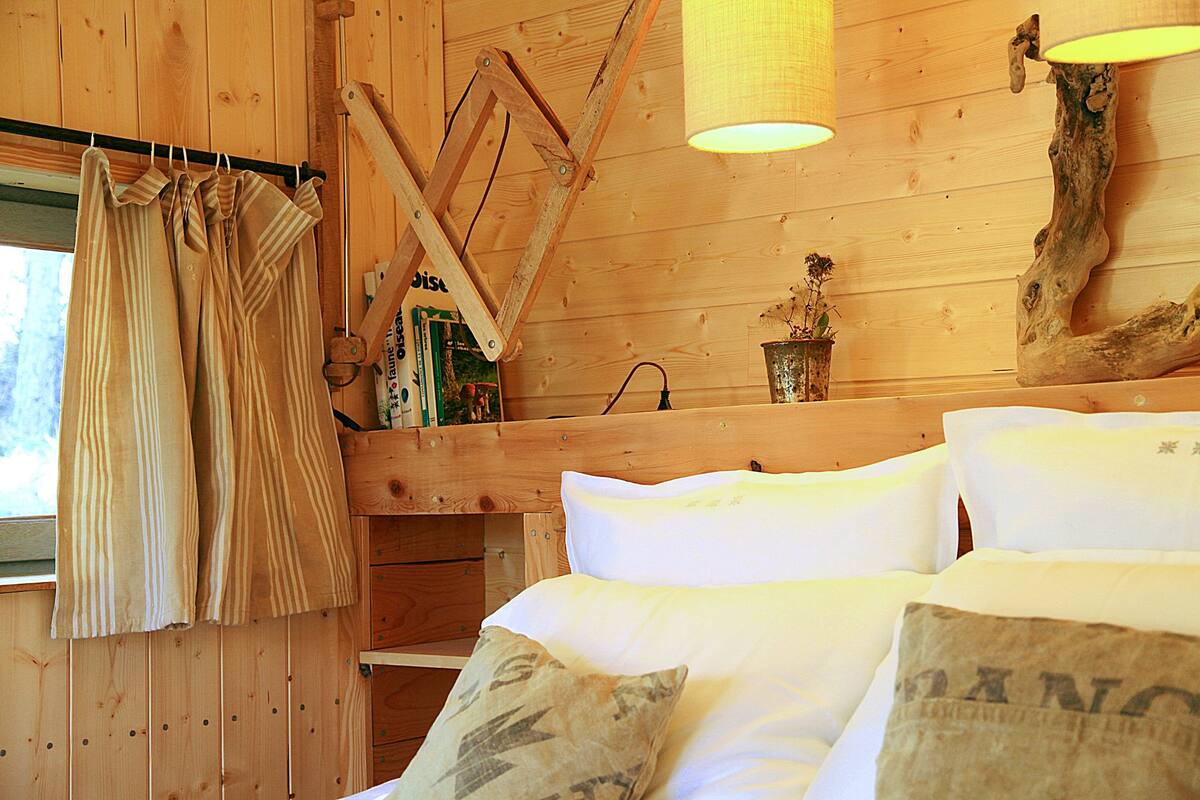 Een houten kamer met een bed, gordijnen, een plank met boeken en een lamp.