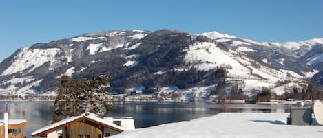 A chalet-style building with a wooden deck and a snow-covered roof, overlooking a lake and mountains.