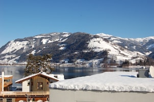 A chalet-style building with a wooden deck and a snow-covered roof, overlooking a lake and mountains.