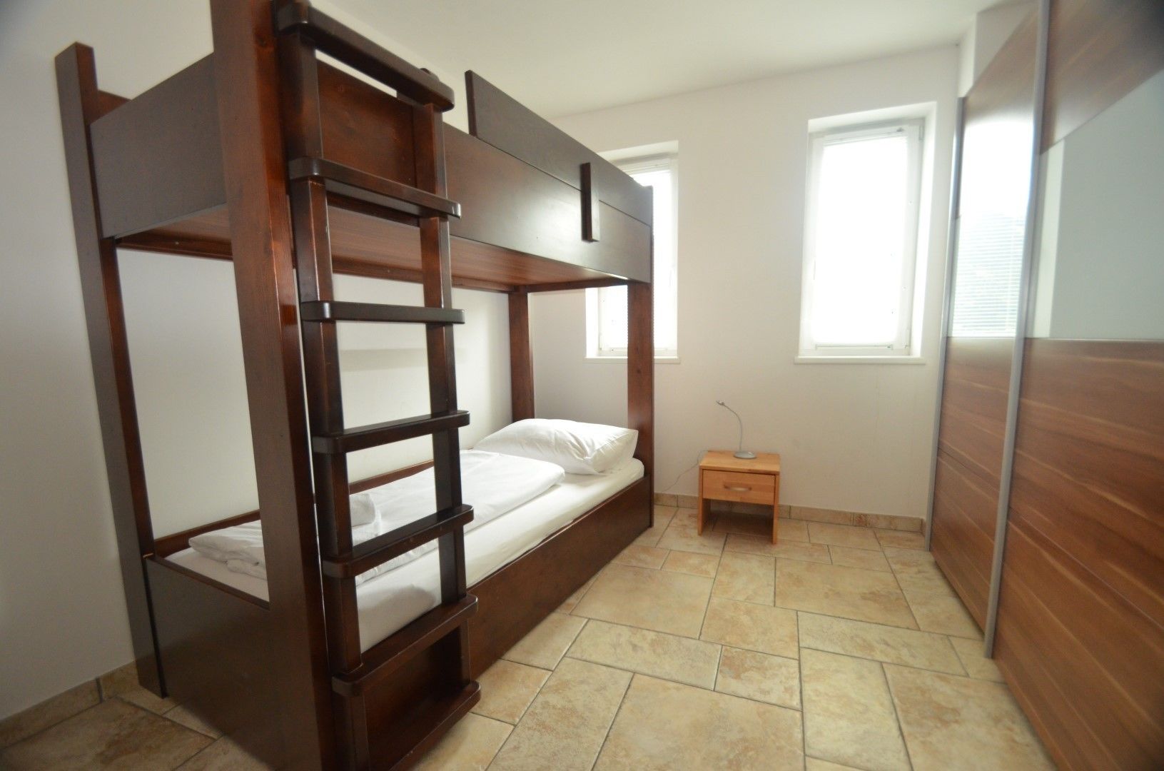 A bunk bed room with a wooden ladder, a bedside table with a lamp, and a window.