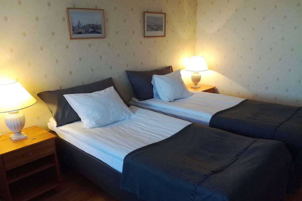 A hotel room with two single beds, each with a pillow and a bedside table with a lamp.