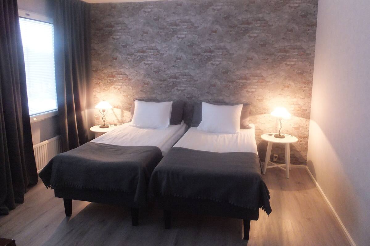 A room with two beds, a brick wall, a window with curtains, and two bedside tables with lamps.
