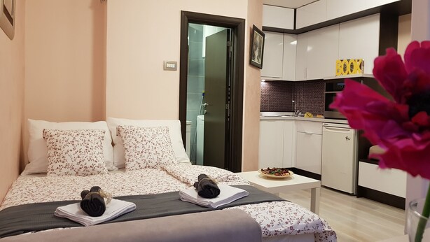 Quarto de hotel moderno com cama, mesa pequena, área de cozinha e banheiro visível por uma porta aberta.