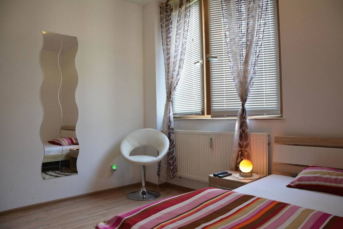 Une chambre à coucher comprenant un lit, une chaise, une table de chevet avec une lampe, une fenêtre avec des stores et un radiateur.