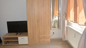 A room with a wooden wardrobe, a small desk with a chair, a TV, and a window with curtains. - Saarbrücken City Apartments (Saarbrücken)